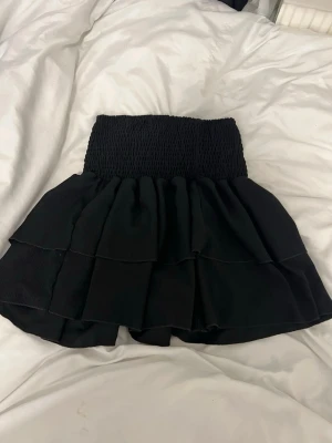 Svart volangkjol med smock - Kort svart kjol med bred smock i midjan och flera lager volanger. Kjolen har en loose passform och är tillverkad i ett mjukt tyg, perfekt för en trendig och bekväm look.