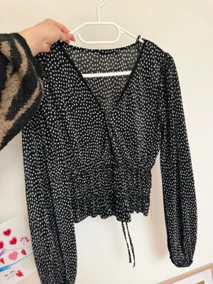 Svart prickig blus från Zara - Snygg svart blus från Zara med vita prickar, v-ringning och smockad midja. Långa puffiga ärmar och knytband i midjan ger en trendig look. Tillverkad i ett lätt och luftigt material som känns skönt mot huden.