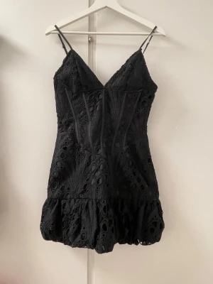 Svart kort klänning med spetsdetaljer från PrettyLittleThing little - Supersnygg svart kort korsettklänning med tunna axelband och v-ringning. Klänningen har ett unikt spetsmönster och volang nedtill som ger en cool och trendig look. Perfekt för dig som vill sticka ut lite extra. Passar till många olika stilar!