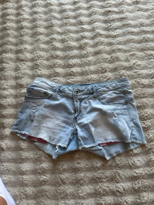 Ljusa jeansshorts med slitningar - Säljer ett par ljusblå jeansshorts med råa kanter och slitna detaljer framtill. Klassisk femficksmodell med knapp och dragkedja. Perfekta för sommaren och ger en avslappnad streetstyle-look.
