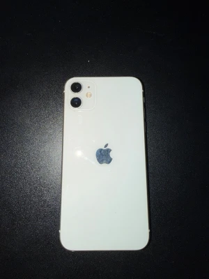 Vit iPhone 11 med dubbla kameror - Snygg vit iPhone 11 med stilren design och rundade hörn. Baksidan är i glas och mobilen har två kameror samt blixt i det övre vänstra hörnet. Apple-loggan sitter i mitten på baksidan. Perfekt för dig som gillar modern teknik och enkel design.