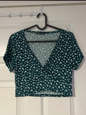 Grön blommig topp - Grön croppad topp med små vita blommor och smickrande omlott-ringning. Korta ärmar och figurnära passform som ger en feminin silhuett. Perfekt för en avslappnad men ändå snygg look lika fin till jeans som till kjol (ifrån shein)  