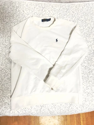Vit sweatshirt från Polo Ralph Lauren Slim Fit - Klassisk vit sweatshirt från Polo Ralph Lauren med rund halsringning och den ikoniska lilla broderade loggan på bröstet. Tröjan har långa ärmar och ribbade muddar vid ärmslut och nederkant. Perfekt för en clean och stilren look.