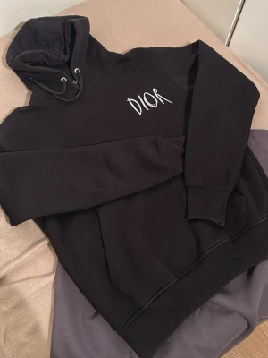 Di*r Svart hoodie - Säljer en svart hoodie från Di*r med vit broderad logga på bröstet. Och fin CD text på ryggen. en stor magficka. Tillverkad i mjukt material som känns skönt mot huden. 
