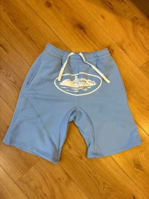 Corteiz shorts  - Shortsen har lite fläckor i bild 3 och 4 så kan man se vart fläckarna är.                 Säljer Corteiz shorts som är ljusa blå shorts med vit snörning i midjan och ett vitt tryck framtill. Shortsen har sidofickor och en bakficka, samt elastisk midja för skön passform. Perfekta för sommaren och avslappnade dagar.
