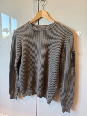 Grå ribbstickad tröja med ficka Lyle & Scott  - Säljer en grå ribbstickad tröja med rund hals och lång ärm. Tröjan har en svart dragkedjeficka på ena ärmen som ger en cool detalj. Perfekt för dig som gillar enkel men snygg stil. Materialet ser ut att vara bomull eller en bomullsblandning.