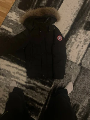 Svart jacka från Canada Goose - Säljer en svart jacka från Canada Goose med avtagbar huva och snygg pälskant. Jackan har klassisk logga på ärmen och stängs med dragkedja och knappar. Perfekt för kalla vinterdagar och riktigt varm.pris kan diskuteras 