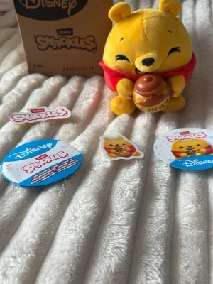 Disney Zuru Snapples Nalle Puh mjukdjur - Supergulligt mjukdjur av Nalle Puh från Disney Zuru Snapples. Gosedjuret är gult med röd tröja och håller en brun honungsburk med texten 'Hunny'. Perfekt som samlarpryl eller present till Disneyfans. 💛