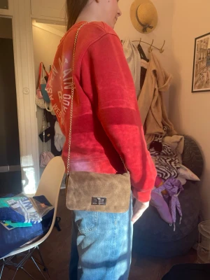 Brun axelväska i mocka med kedja - Snygg brun axelväska i mocka-look med guldfärgad kedjerem och silverfärgat vridlås. Väskan har en mjuk, rektangulär form och passar perfekt till jeans eller en chill outfit. Enkel men trendig design som ger en stilren vibe.