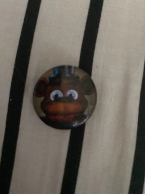 Svart katt-pin med tecknad design - Säljer en rund pin med en svart tecknad katt på grå bakgrund. Katten har stora vita ögon och en lite sarkastisk blick. Perfekt att fästa på väskan eller jackan för en personlig touch.