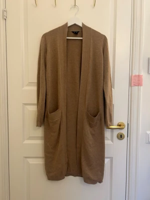 Beige lång kofta med fickor - Lång, öppen beige kofta med två stora fickor framtill. Koftan har en rak passform och långa ärmar, perfekt att slänga över en outfit för extra lager. Enkel och stilren design utan knappar eller dragkedja.