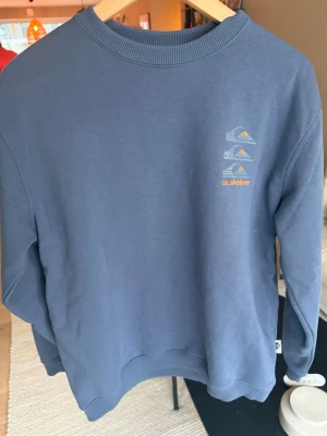 Blå sweatshirt från Quiksilver - Säljer en blå sweatshirt från Quiksilver med tryck av vågor och berg i vitt och orange på bröstet. Tröjan har rund halsringning, ribbade muddar och är gjord i mjukt bomullsmaterial. Perfekt för en avslappnad och stilren look.