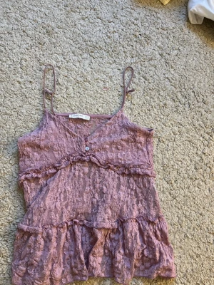 Ljuslila spetslinnetopp - Säljer en ljuslila linnetopp från Stradivarius med tunna axelband och v-ringning. Toppen är i spets med volangdetaljer och en liten knapp framtill. Perfekt för sommarens alla outfits! Liten i storleken💖