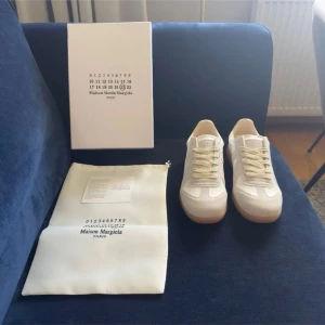 Maison Margiela vita sneakers - Snygga vita sneakers från Maison Margiela med beige detaljer och klassisk gummisula. Skorna har en stilren design med ovandel i läder och mocka, samt vita skosnören. Perfekta för dig som gillar minimalistisk och trendig stil.