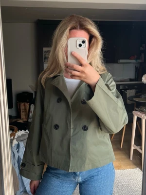 Kort grön trenchcoat bYIC - Stilren kort trenchcoat i olivgrönt med dubbelknäppning framtill och klassisk krage. Jackan har två snedställda fickor och dekorativa knappar vid ärmsluten. Storlek S BYIC.