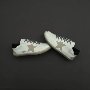 Vita golden goose sneakers - Snygga vita sneakers från golden goose med beige stjärna på sidan och svart hälparti. Skorna har gråa snören och en klassisk låg siluett. Perfekta för dig som gillar stilrena detaljer och en modern look.