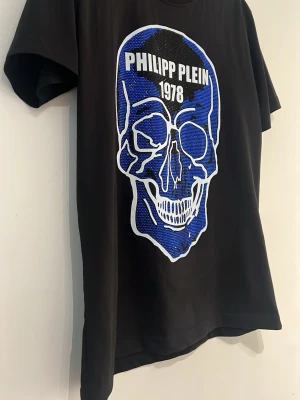 Svart Philipp Plein t-shirt med dödskalle - Cool svart t-shirt från Philipp Plein med stor blå och vit dödskalle framtill och texten 'PHILIPP PLEIN 1978' ovanför motivet. T-shirten har klassisk passform och korta ärmar. Perfekt för dig som gillar statement-plagg.
