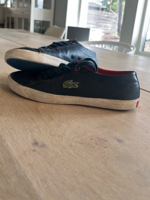 Marinblåa Lacoste sneakers i läder - Snygga marinblå sneakers från Lacoste med klassisk krokodillogo på sidan. Skorna har vita sulor, svarta skosnören och röda detaljer på insidan. Tillverkade i läder med textilfoder för en clean och stilren look.