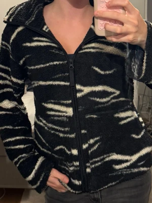Svart teddyjacka med zebramönster - Mysig svart teddyjacka med vitt zebramönster och dragkedja framtill. Jackan har hög krage, långa ärmar och två fickor på sidorna. Perfekt för dig som vill ha en trendig och bekväm look med lite edge.
