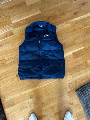 Mörkblå dunväst från Nike - Snygg mörkblå dunväst från Nike med dragkedja framtill och två stora fickor. Västen har hög krage och klassisk Nike-logga på bröstet. Perfekt för lager-på-lager och streetwear-stil. Materialet är glansigt och quiltat för extra värme.