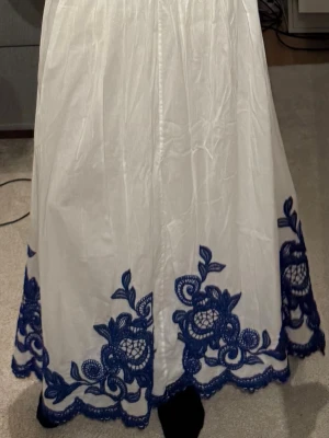 Vit långkjol med blå broderi - Vit långkjol med vackra blå broderade blommor längs nederkanten. Kjolen har en luftig och elegant form, perfekt för dig som vill sticka ut med en unik detalj. Materialet känns lätt och passar bra till vår och sommar.