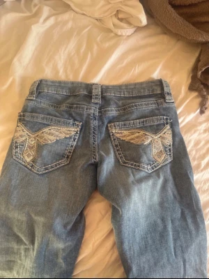 Blå jeans med broderade bakfickor - Säljer ett par ljusblå jeans med snygga broderade detaljer i vitt och beige på bakfickorna. Jeansen har normal midja, bälteshällor och klassisk femficksmodell. Perfekta för dig som vill ha något unikt och trendigt i garderoben. Strl 158~xs, hör av dig vid frågor!🫶😊