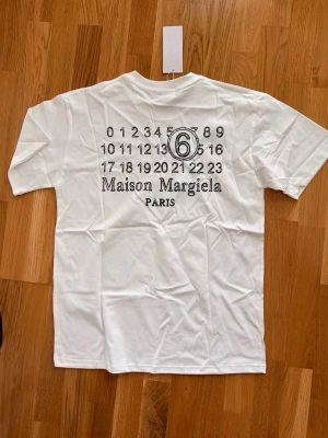 Maison Margiela t-shirt - Vit t-shirt från Maison Margiela med tryck på ryggen och diskreta sömmar framtill. Klassisk passform och rund halsringning. Perfekt för dig som gillar stilrena och trendiga plagg med en twist.