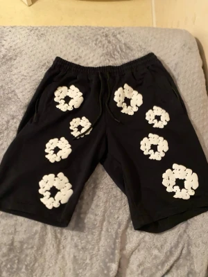 Svarta shorts med vita blommor - Säljer ett par svarta shorts med elastisk midja och snörning. Shortsen har stora vita blommönster tryckta över hela plagget, vilket ger en unik och trendig look. Perfekta för dig som vill sticka ut i sommarvärmen.