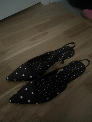 Svarta prickiga slingback-pumps - Snygga svarta pumps med vita prickar och spetsig tå. Skorna har slingback-rem och låg klack. Helt nya och oanvända. 