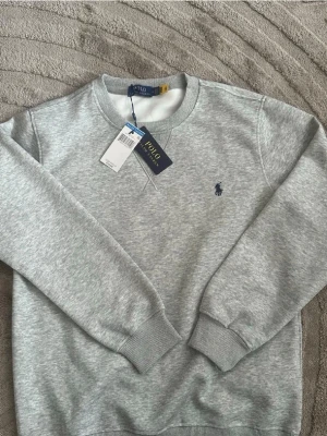Grå sweatshirt från Polo Ralph Lauren - Grå sweatshirt från Polo Ralph Lauren med rund halsringning och broderad marinblå logga på bröstet. Tröjan har ribbade muddar vid ärmslut och nederkant, perfekt för en avslappnad och stilren look.