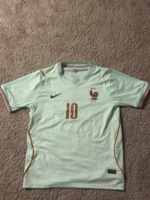 Frankrike VM borta tröja Mbappe 2026 - Helt ny fotbollströja från Nike med Frankrikes landslagsemblem och nummer 10 i guld på bröstet. På ryggen står det Mbappé och nummer 10. 