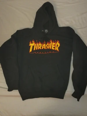 Svart Thrasher hoodie med flames - Svart hoodie från Thrasher med den ikoniska loggan i gult och rött flames-tryck på bröstet. Perfekt för dig som gillar streetwear och skatekultur.