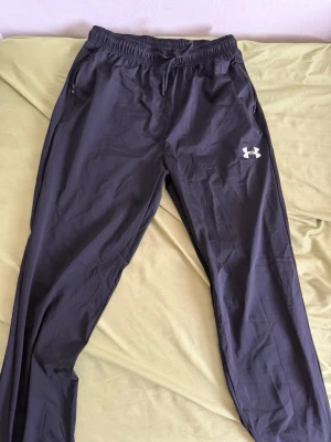 Svarta mjukisbyxor från Under Armour - Säljer ett par svarta mjukisbyxor från Under Armour med resår i midjan och dragsko. Byxorna har dragkedjeförsedda fickor och logga på vänster lår. Tillverkade i ett lätt och sportigt material, perfekta för chill eller träning.