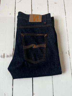 Mörkblå jeans från Nudie Jeans - Säljer ett par mörkblå jeans från Nudie Jeans med klassisk femficksmodell och orange kontrastsömmar. Jeansen har rak passform och är tillverkade i kraftig denim. Snygg detalj med vågig söm på bakfickan och läderpatch i midjan.