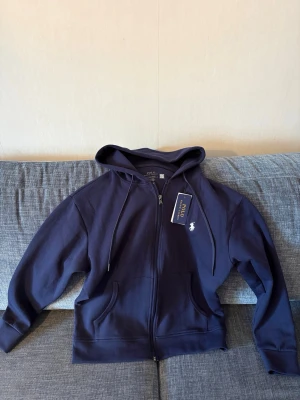 Mörkblå hoodie från Polo Ralph Lauren - Snygg mörkblå hoodie från Polo Ralph Lauren med dragkedja framtill, två fickor och klassisk huva. Ikoniska loggan broderad på bröstet. Tillverkad i mjukt bomullsmaterial som känns skönt mot huden. Perfekt för en chill och stilren look.