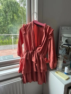 Rosa volangklänning med v-ringning - Säljer en kort rosa klänning med långa ärmar och djup v-ringning. Klänningen har flera volanglager och en omlott-look framtill. Perfekt för dig som gillar feminina detaljer och en trendig stil.