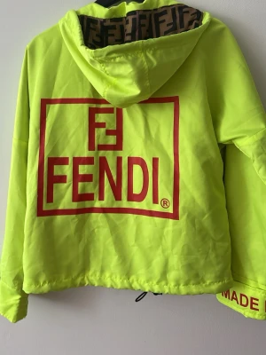 Neongul vindjacka Fendi - Neongul vindjacka Fendi med stor röd logga på ryggen och textdetaljer på ärmarna. Jackan har huva med Fendi-mönstrat foder, svarta dragkedjor och justerbar nederkant. Perfekt för dig som vill sticka ut med en exklusiv streetwear-look. Kom gärna med prisförslag!