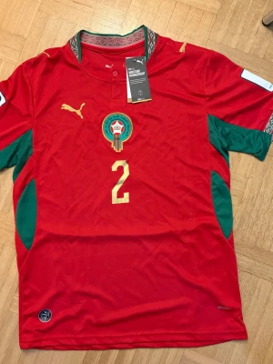 Marocko fotbollströja Hakimi Puma - Säljer en röd Marocko landslagströja från Puma med Hakimi och nummer 2 på ryggen. Tröjan har gröna detaljer på sidorna och ärmarna samt guldfärgade logotyper och siffror. Tillverkad i ett lätt och ventilerande material, perfekt för fotbollsträning eller match.