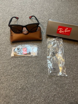 Svarta Ray-Ban solglasögon Wayfarer - Säljer ett par klassiska svarta Ray-Ban Wayfarer solglasögon med mörka glas och röda detaljer på skalmarna. Kommer med originalfodral, putsduk och box. Perfekta för dig som vill ha en tidlös och snygg look i solen.