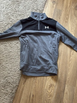 Grå träningsjacka från Under Armour - Snygg grå träningsjacka från Under Armour med svart panel upptill och halv dragkedja. Jackan har hög krage, två sidofickor med dragkedja och logga på bröstet. Perfekt för träning eller chill. Materialet är mjukt och stretchigt för bästa komfort.