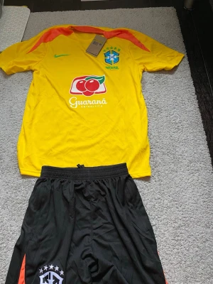 Brasilien fotbollströja och shorts - Säljer ett set med Brasilien fotbollströja i gult med gröna och orange detaljer samt matchande svarta shorts. Tröjan har CBF-logga, Nike-logga och Guaraná Antarctica-tryck. Perfekt för fotbollsträning eller match.