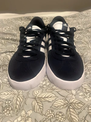 Mörkblåa Adidas sneakers med vita ränder - Snygga svarta Adidas sneakers med klassiska vita ränder på sidorna och vit sula. Skorna har lågt skaft, svarta skosnören och ovandel i mockaliknande material. Perfekta för en stilren och sportig look.