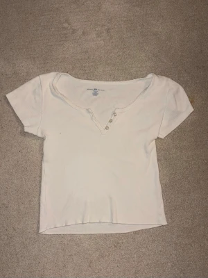 Vit ribbad topp med knappar från brandy Melville  - Vit ribbad topp med korta ärmar och v-ringning med tre knappar framtill. Enkel och stilren design som passar perfekt till jeans eller kjol. Mjuk och stretchig känsla.