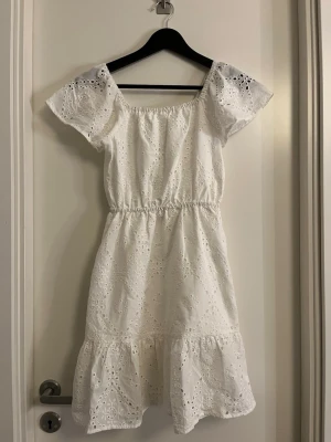 Vit broderad klänning från Lindex - Söt vit klänning från Lindex med broderade hålmönster och resår i midjan.  Klänningen har korta ärmar och volang nedtill, perfekt för sommarens alla tillfällen. Tillverkad i ett lätt och luftigt material. Endast använd några få gånger. 