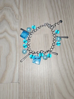 Armband med blå pärlor och berlocker - Charmigt armband med silverfärgad kedja, dekorerat med blåa runda och fyrkantiga pärlor samt silvriga stavar. Pärlorna är i olika nyanser av blått och ger ett lekfullt och trendigt intryck. Perfekt accessoar för att lyfta din outfit.