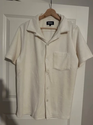 Beige frottéskjorta från Nikben - Kortärmad skjorta från Nikben i mjuk vit frotté. Klassisk krage, knappstängning framtill och en bröstficka. Perfekt för en chill och avslappnad stil.