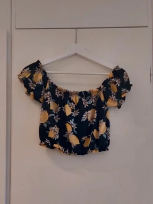 Blommig offshoulder topp i mörkblått - Söt offshoulder topp med blommigt mönster i gult och vitt på mörkblå botten. Kort och croppad modell med resår vid ärmslut och midja. Perfekt för sommarens alla tillfällen!