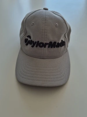 Grå keps från TaylorMade - Snygg grå keps från TaylorMade med böjd skärm och svart broderad logga framtill. Kepsen har klassisk sexpanelsdesign och justerbar rem bak. Tillverkad i slitstarkt tyg som passar perfekt till en sportig look.