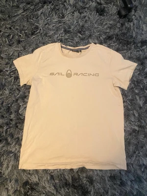 Beige Sail Racing t-shirt - Säljer en beige t-shirt från Sail Racing med tryck på bröstet och en liten logga i nacken. Klassisk rund hals och korta ärmar. Perfekt för dig som gillar enkel och stilren design.(äkta)