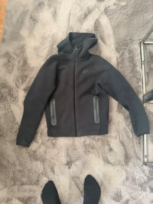 Svart Nike hoodie med dragkedja - Snygg svart hoodie från Nike med hel dragkedja framtill och två stora fickor med dragkedja. Hoodien har en klassisk huva och diskret Nike-logga på bröstet. Tillverkad i ett mjukt och tjockt material som passar perfekt till chill eller streetwear.